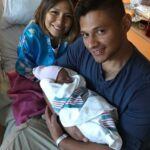 A woman in a hospital gown looks over the shoulder of a man as he cradles an infant in his arms, representing the joy of new parents in the US military.