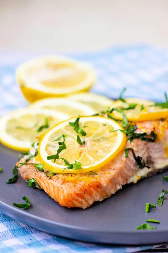 A dark plate featuring a piece of beautifully baked salmon, garnished with lemon slices and fresh herbs, highlighting its appetizing appearance and flaky texture.