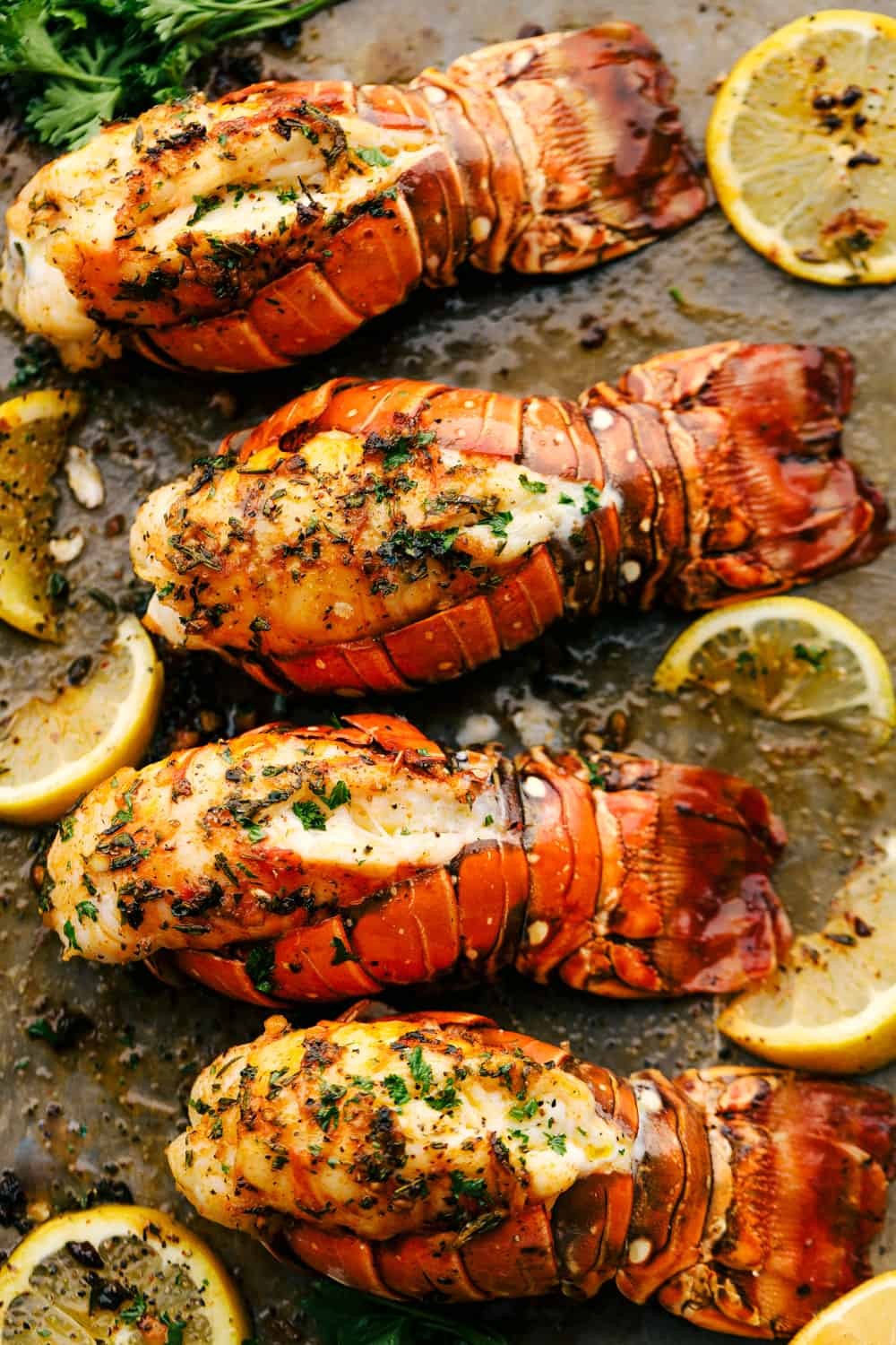 Broiled lobster tails with a golden-brown garlic herb butter topping, served on a baking sheet.