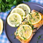 A plate containing a piece of perfectly cooked salmon topped with vibrant lemon slices and fresh green herbs, showcasing its flaky texture and moist appearance.