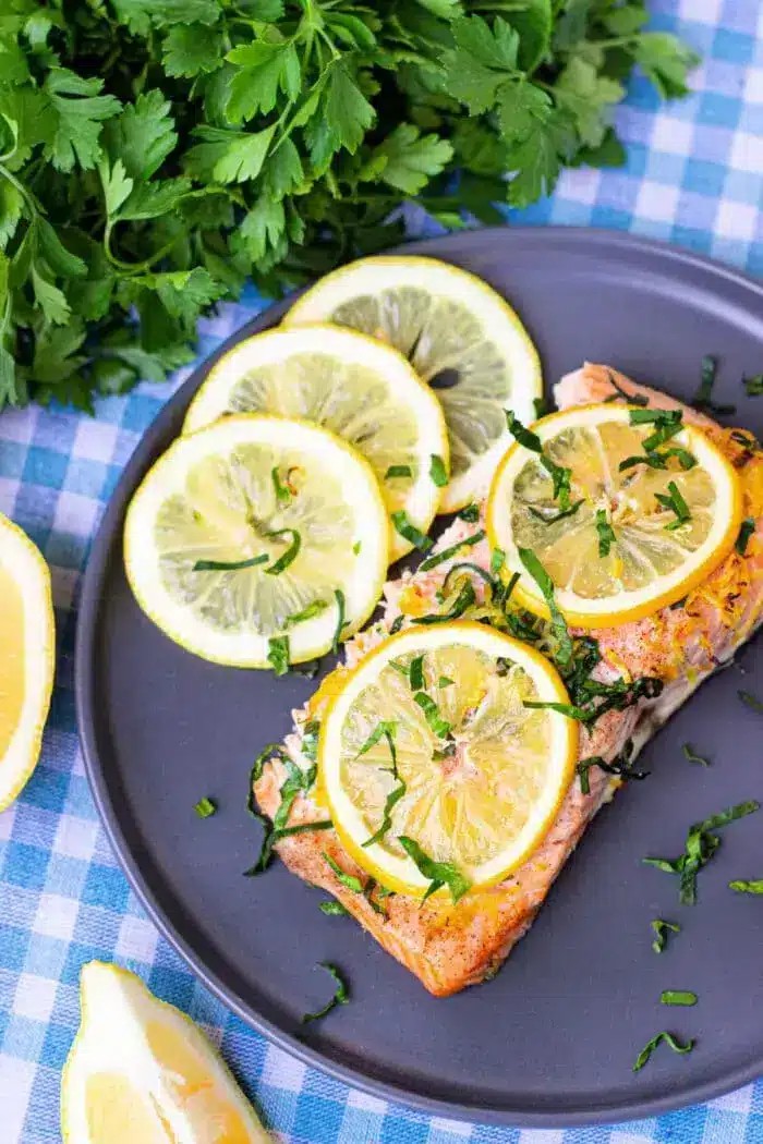 A plate containing a piece of perfectly cooked salmon topped with vibrant lemon slices and fresh green herbs, showcasing its flaky texture and moist appearance.