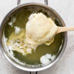 A close-up shot of a spoon lifting a perfectly boiled chicken thigh from a pot of simmering water.