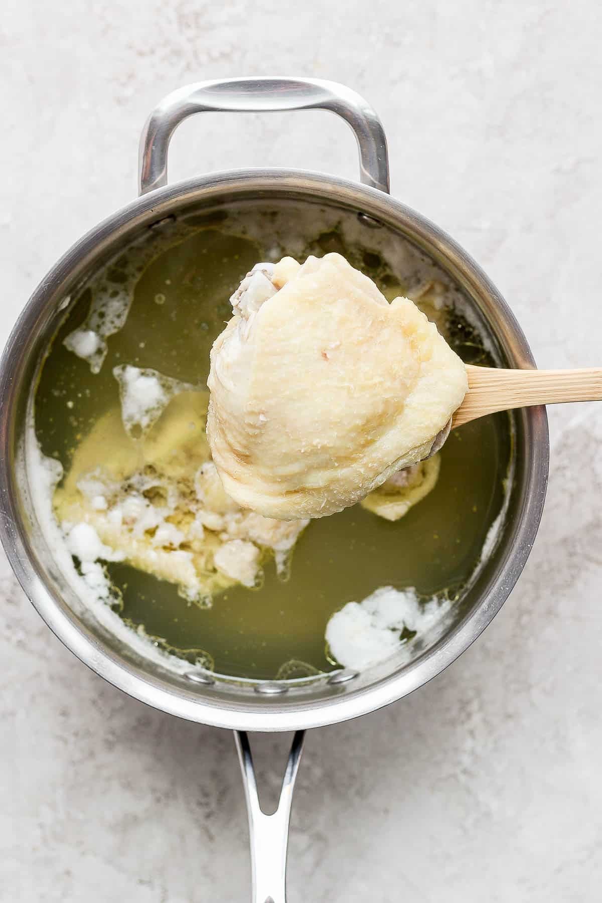 A close-up shot of a spoon lifting a perfectly boiled chicken thigh from a pot of simmering water.