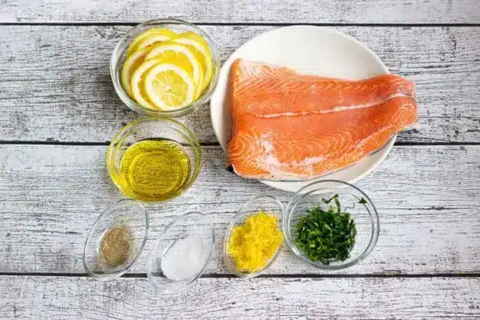 A close-up shot of raw salmon fillets on a white surface, surrounded by small bowls of ingredients like lemon slices, olive oil, herbs, salt, and pepper, ready for seasoning and baking.