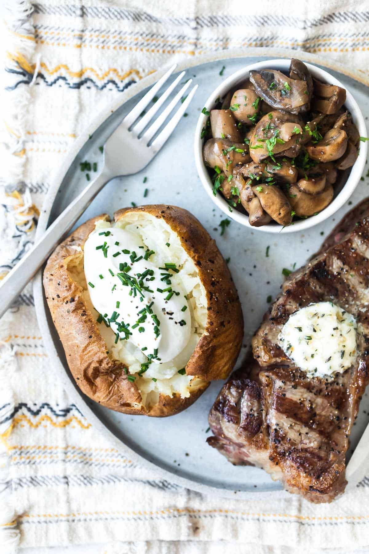 A plate with a baked potato topped with sour cream, a steak topped with butter, and a bowl of balsamic mushrooms on a plate.