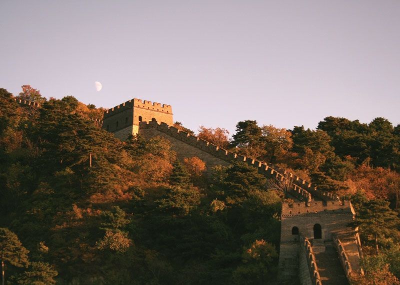 Mountainous landscape with the Great Wall of China winding across the peaks, under a rising moon.