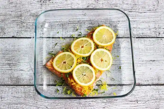 A glass baking dish showcasing a perfectly baked salmon fillet, topped with bright yellow lemon slices and fresh green herbs, ready to be served.