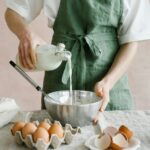 Woman pouring milk from a glass bottle into the bowl with eggs in front.