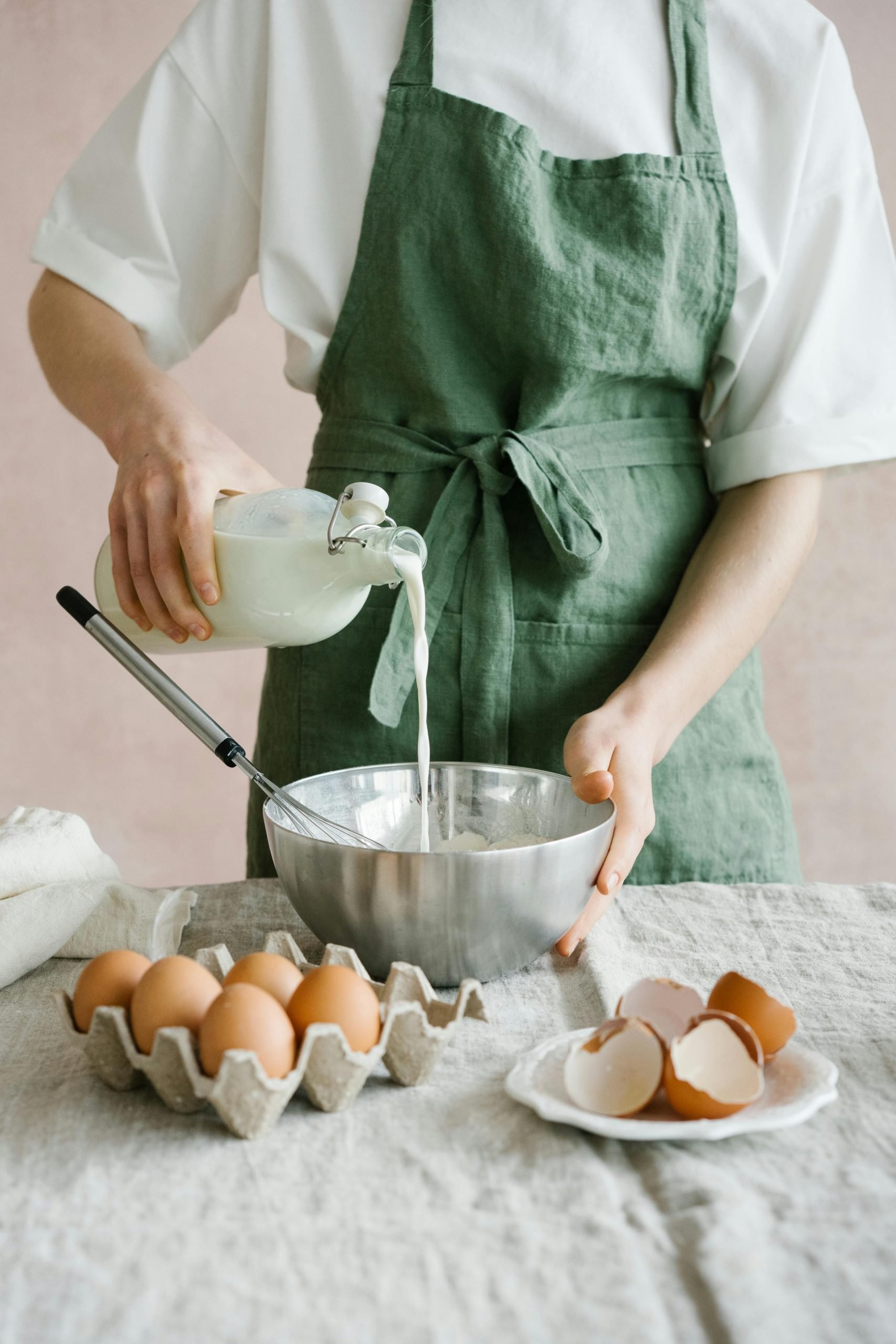 Woman pouring milk from a glass bottle into the bowl with eggs in front.