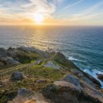 Four white-sided, red-roofed structures sit on a rocky headland above the Pacific Ocean at the base of a long stairway. Above and to the left of the lighthouse, the sun filters through wispy clouds as it descends toward the horizon.