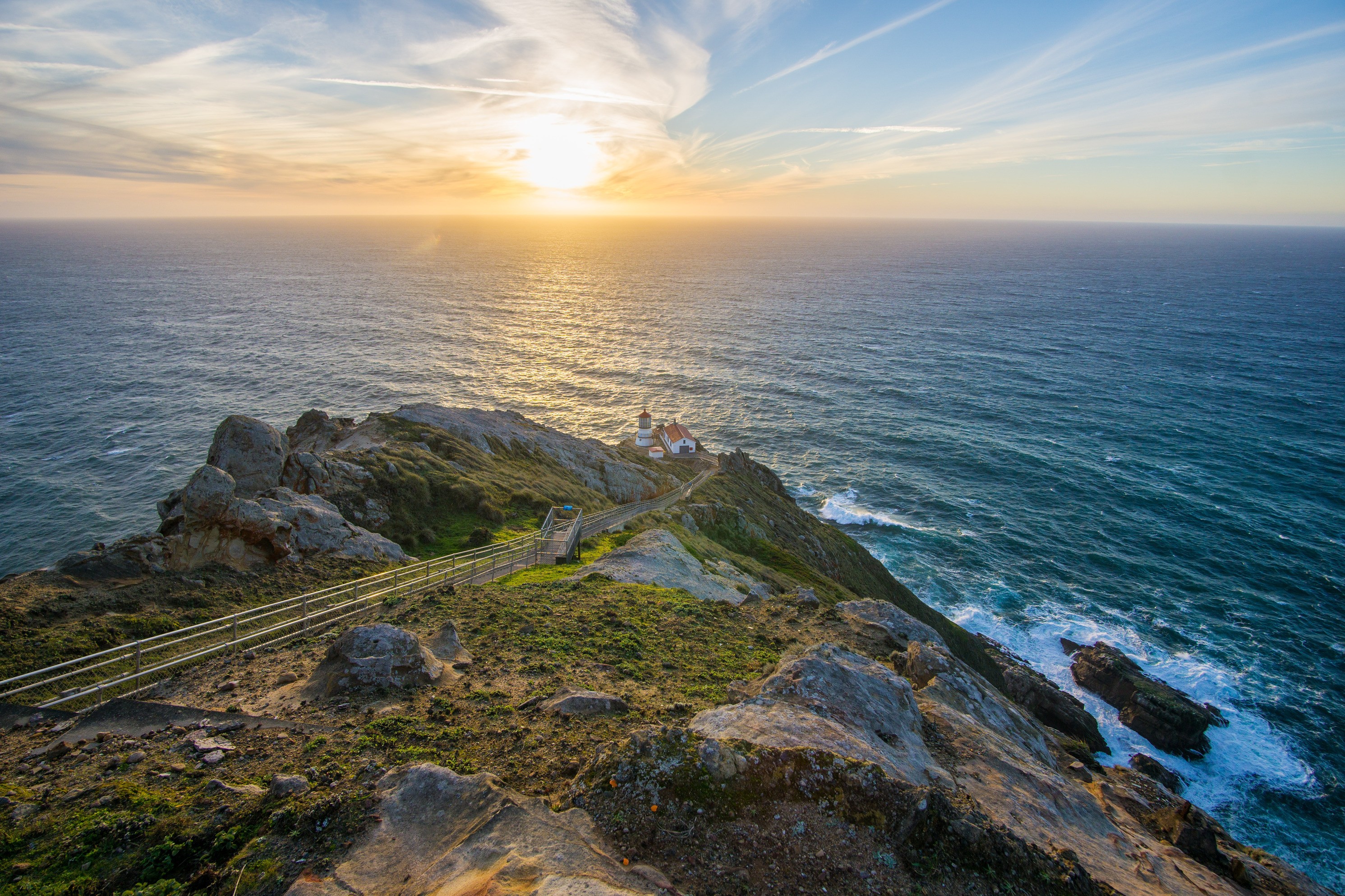 Four white-sided, red-roofed structures sit on a rocky headland above the Pacific Ocean at the base of a long stairway. Above and to the left of the lighthouse, the sun filters through wispy clouds as it descends toward the horizon.