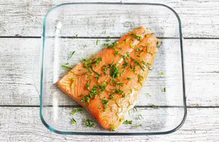 A close up image of a raw salmon fillet, seasoned generously with freshly chopped green herbs, ready to be baked.