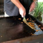 Scraping food debris from a hot Blackstone griddle using a metal spatula