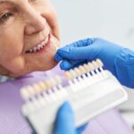 Gloved left hand of dentist holding implant tray and right hand holding implant to woman's mouth, illustrating implant-supported dentures procedure