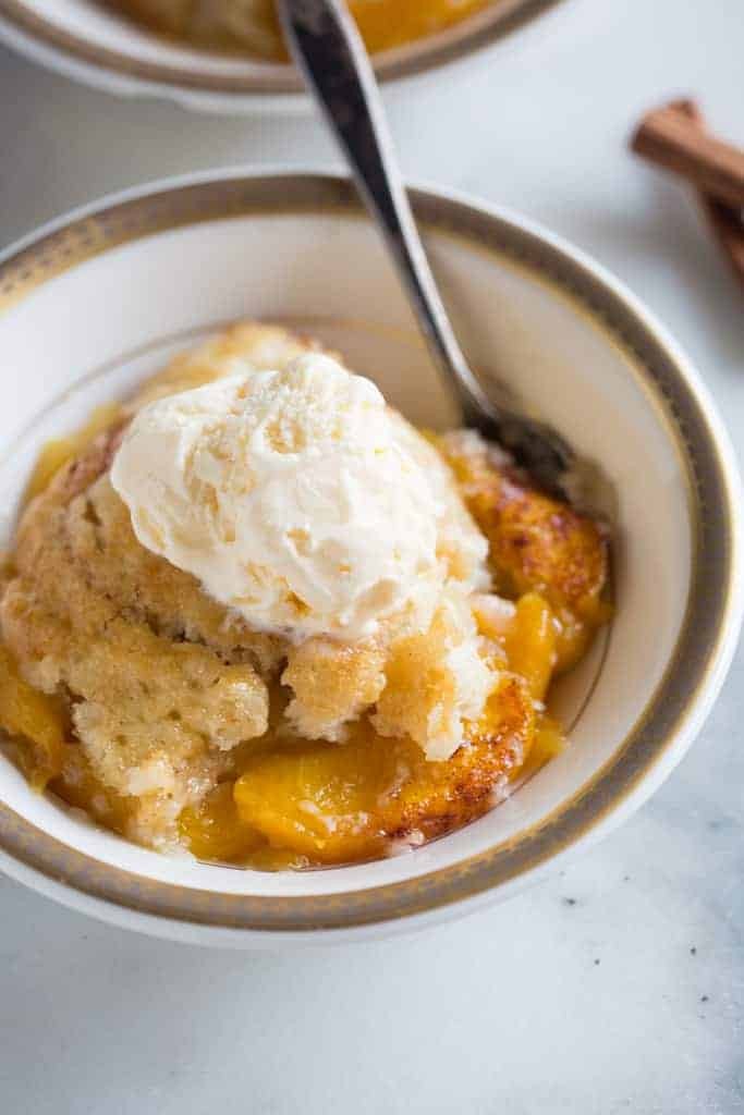 Close-up of a slice of peach cobbler on a white plate, topped with a scoop of vanilla ice cream and a sprig of mint.