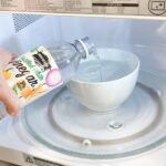 A person pouring vinegar from the bottle in a bowl of water that is sitting in a microwave.
