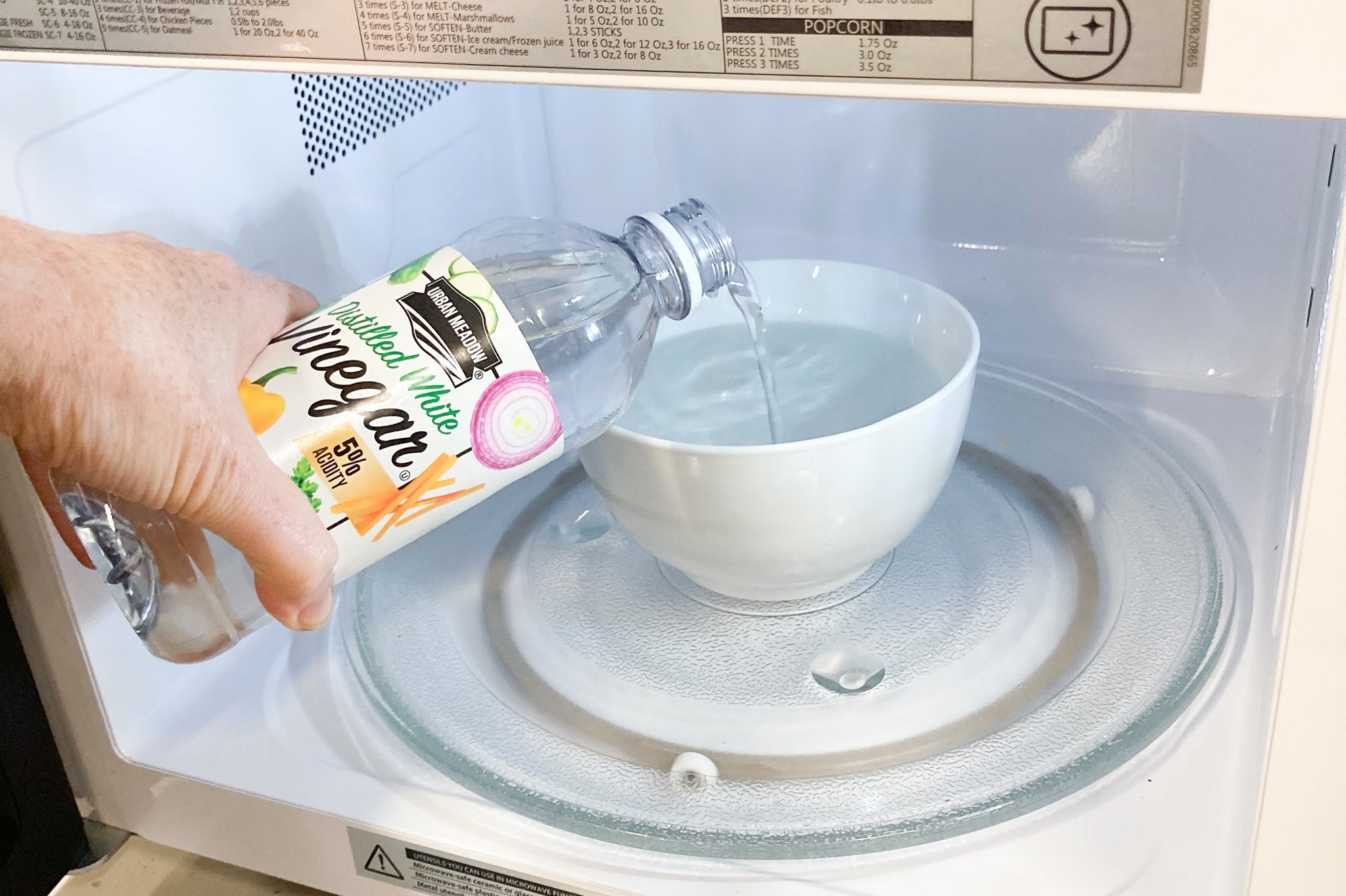 A person pouring vinegar from the bottle in a bowl of water that is sitting in a microwave.