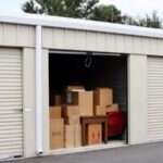 Exterior view of several side-by-side outdoor self-storage units that are painted beige. The center unit has its door open. Inside is some furniture and many cardboard boxes.