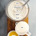 Ingredients for drop dumplings arranged in bowls on a cutting board. From top to bottom: flour, baking powder, salt, pepper, thyme, milk and butter.