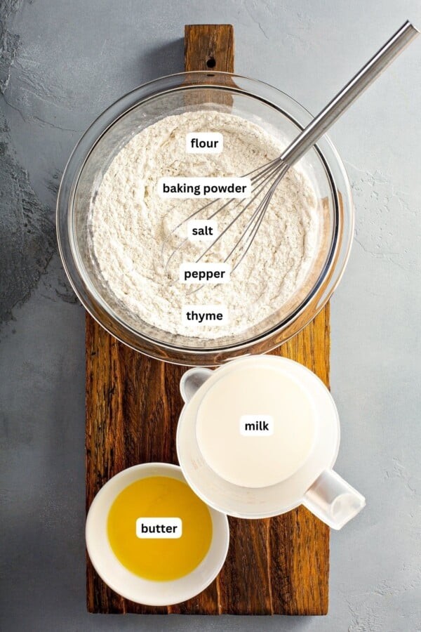 Ingredients for drop dumplings arranged in bowls on a cutting board. From top to bottom: flour, baking powder, salt, pepper, thyme, milk and butter.
