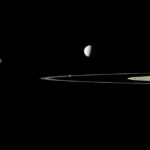 Against a black background, five golden-gray moons of varying sizes are scattered along a horizontal band through the image. Sunlight illuminates them from the left, and their right sides are in shadow. The largest, at far right, takes up the middle third of the photo, fully sunlit, with its dark half cut off by the right edge of the frame. The rings of Saturn, seen nearly edge-on, pierce the image like a knife blade from center-right, emerging through the largest moon, and with the smallest moon resting atop the blade like a breadcrumb.