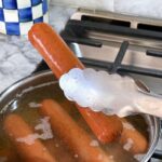 A hot dog is being lifted from a pot of boiling water with tongs.