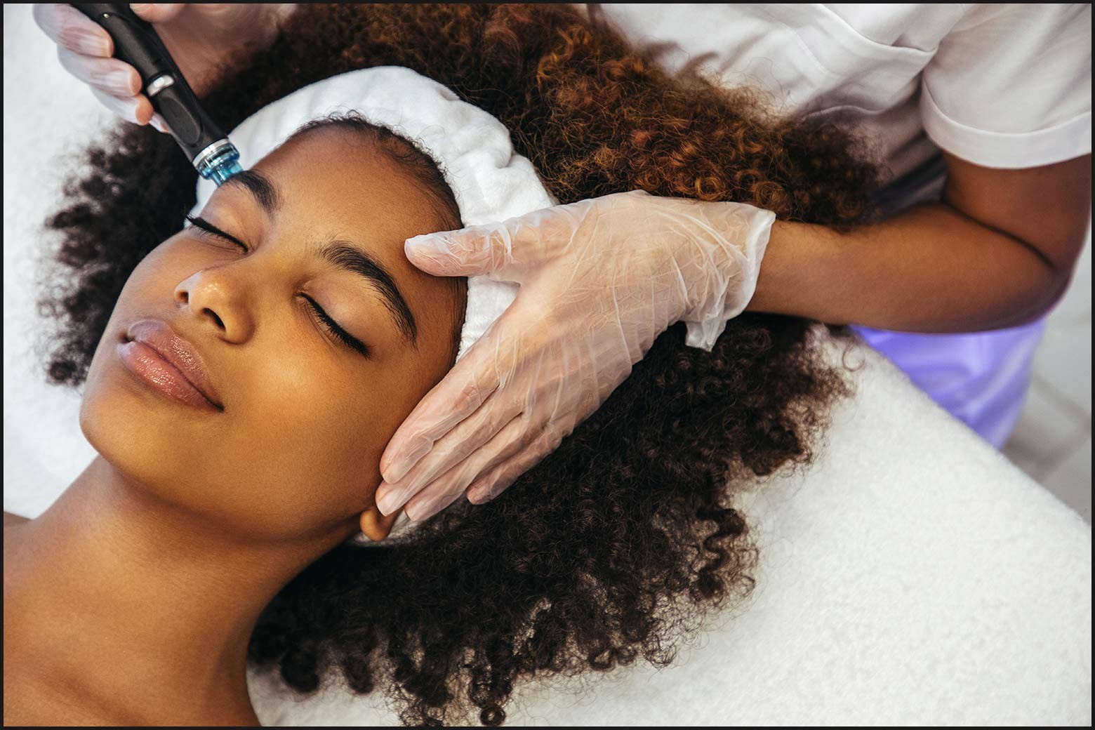 Close up of person’s face with eyes closed and natural hair pulled back, getting a microdermabrasion facial, gloved hand is holding one side of their face
