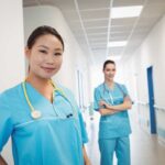 Two nurses in a hospital hallway wearing blue scrubs.
