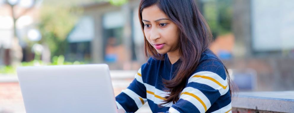 South Asian American student focused on her laptop while applying to college in an outdoor park setting
